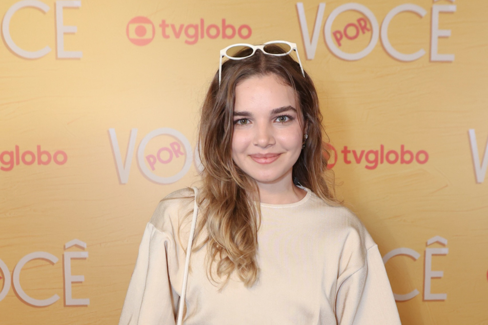 Mulher jovem de cabelos castanhos ondulados, com óculos brancos na cabeça, sorri levemente para a câmera. Veste blusa bege de mangas longas e está em frente a um fundo amarelo com o logo tvglobo e a palavra VOCÊ em branco e rosa
