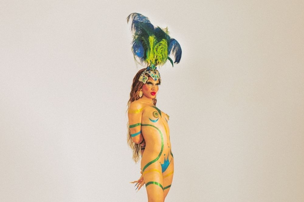Pabllo Vittar posa de perfil, com maquiagem e body painting em tons de verde, azul e amarelo, simulando a bandeira brasileira. Ela usa um cocar de penas azuis e verdes, e tem os cabelos longos e castanhos soltos. As mãos estão amarradas atrás das costas, e ela tem unhas postiças longas e douradas