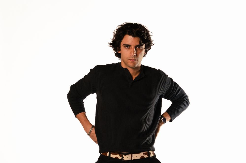 Homem jovem de pele clara, cabelo cacheado escuro, vestindo suéter preto e cinto estampado de vaca, mãos na cintura, olhando sério para frente, em fundo branco