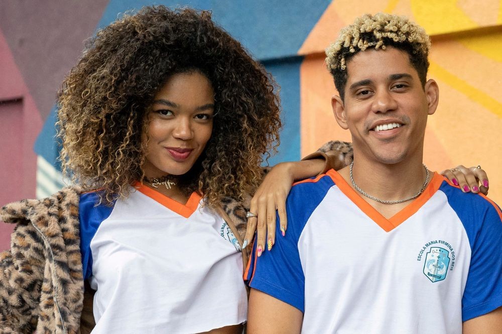 Jovem mulher negra com cabelo cacheado e jaqueta de oncinha, sorrindo ao lado de um homem negro com cabelo loiro e crespo, ambos vestindo camisetas de uniforme escolar brancas com detalhes azuis e laranjas, e um brasão no peito direito do homem. A mulher tem a mão no ombro do homem, e ambos olham para frente com expressões alegres. O fundo é uma parede colorida com tons de rosa, azul e laranja