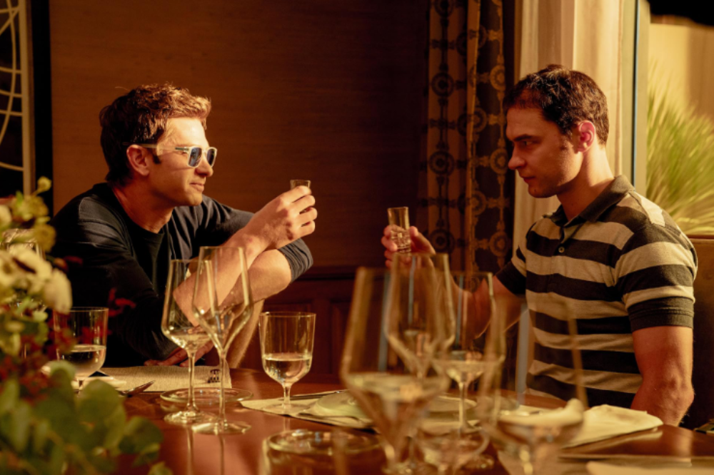 Dois homens sentados à mesa, um de óculos escuros e blusa escura, o outro de camisa polo listrada, brindam com pequenos copos de shot. A mesa está posta com taças e guardanapos, em um ambiente com cortinas estampadas e iluminação quente