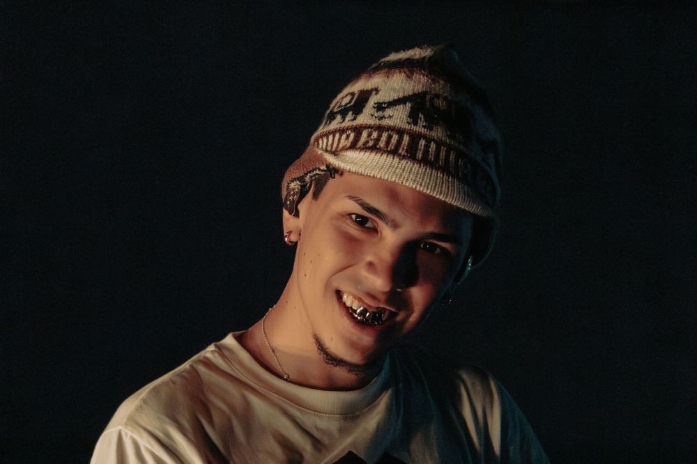 Jovem com boné de lã bege e marrom, sorrindo e mostrando dentes com grillz prateado, em fundo escuro