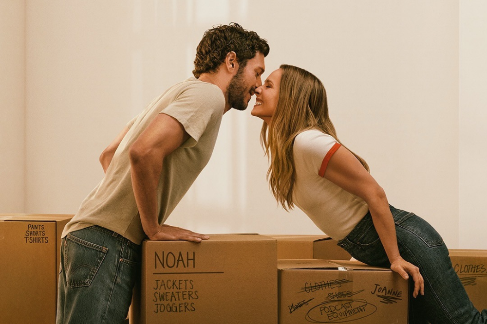 Um casal sorridente, ele com barba e camiseta bege, ela com camiseta branca e jeans, se inclina sobre caixas de mudança, quase se beijando. As caixas têm anotações como Noah, Jackets, Sweaters, Joggers e Joanne, Podcast Equipment