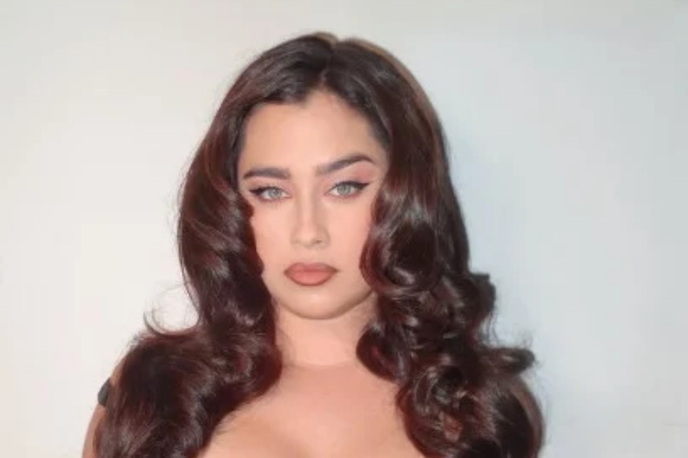 Mulher jovem de pele clara, cabelos castanhos escuros e ondulados, maquiagem marcante nos olhos e lábios, olhando diretamente para frente com expressão séria