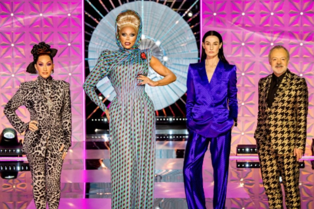 Quatro pessoas posam em um palco iluminado com fundo rosa e azul. Da esquerda para a direita: uma mulher de macacão estampado de onça, RuPaul de vestido longo estampado e turbante, uma mulher de terno roxo brilhante e um homem de terno xadrez preto e dourado