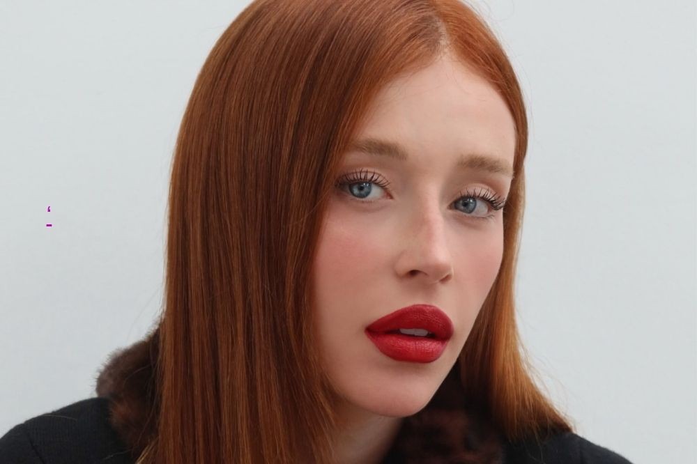 Mulher jovem de cabelos ruivos longos e lisos, olhos azuis claros, maquiagem leve e batom vermelho intenso, olhando diretamente para frente, com um fundo branco