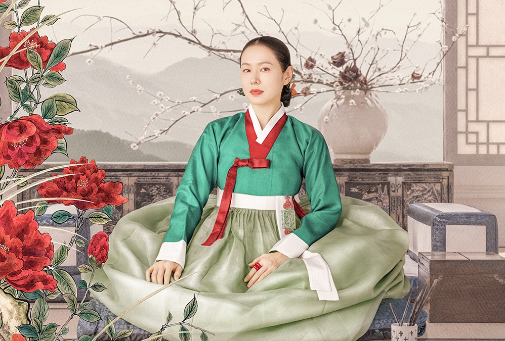 Mulher coreana de pele clara, com cabelo escuro preso, vestindo um hanbok verde-esmeralda e saia verde-clara, com detalhes vermelhos e brancos. Ela está sentada em um cenário tradicional com montanhas ao fundo, flores vermelhas grandes à esquerda e galhos floridos em um vaso à direita