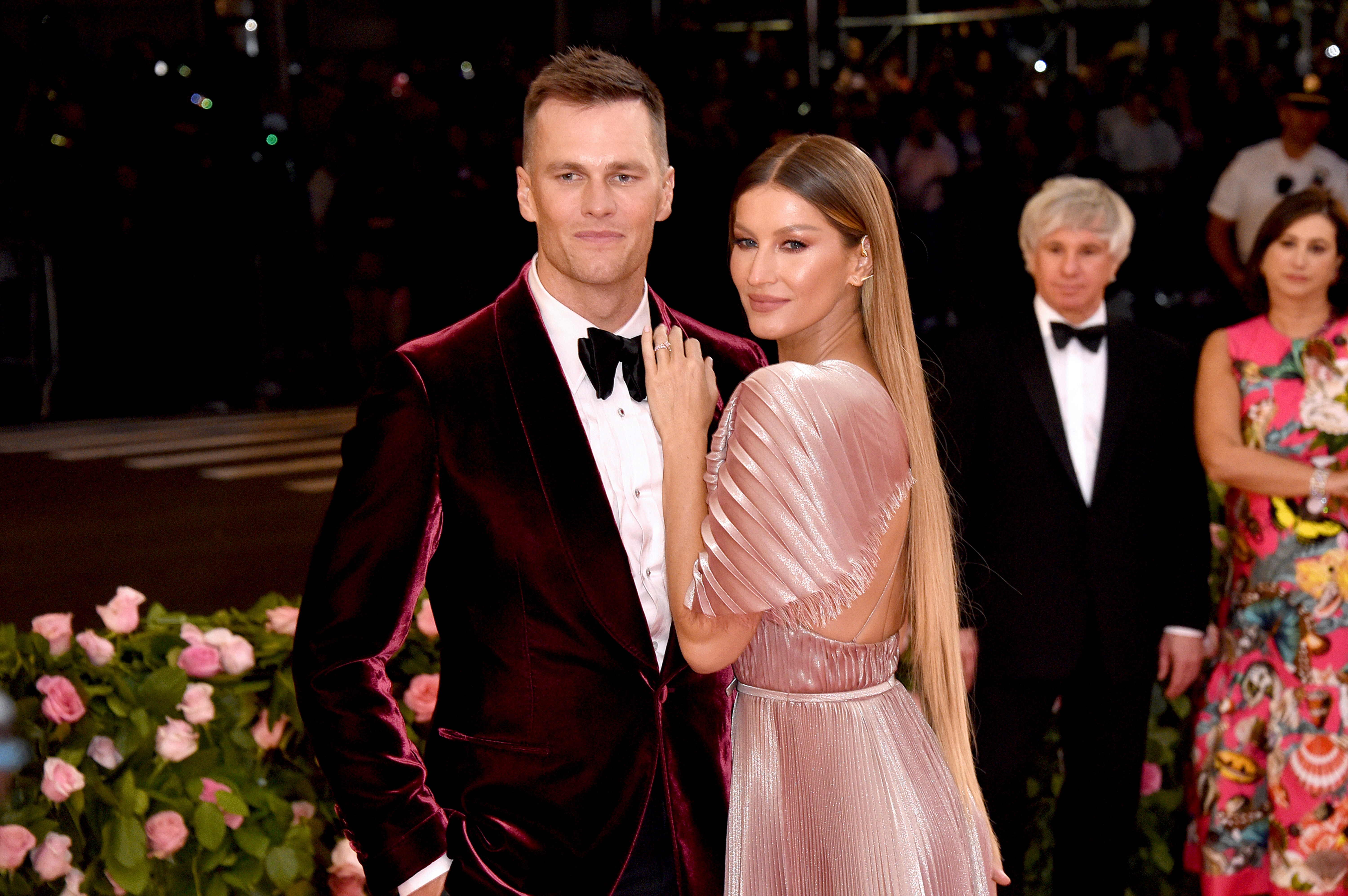 Tom Brady e Gisele Bündchen posam em evento. Ele veste um terno de veludo bordô e gravata borboleta preta. Ela usa um vestido plissado rosa claro com as costas abertas e cabelo longo solto.