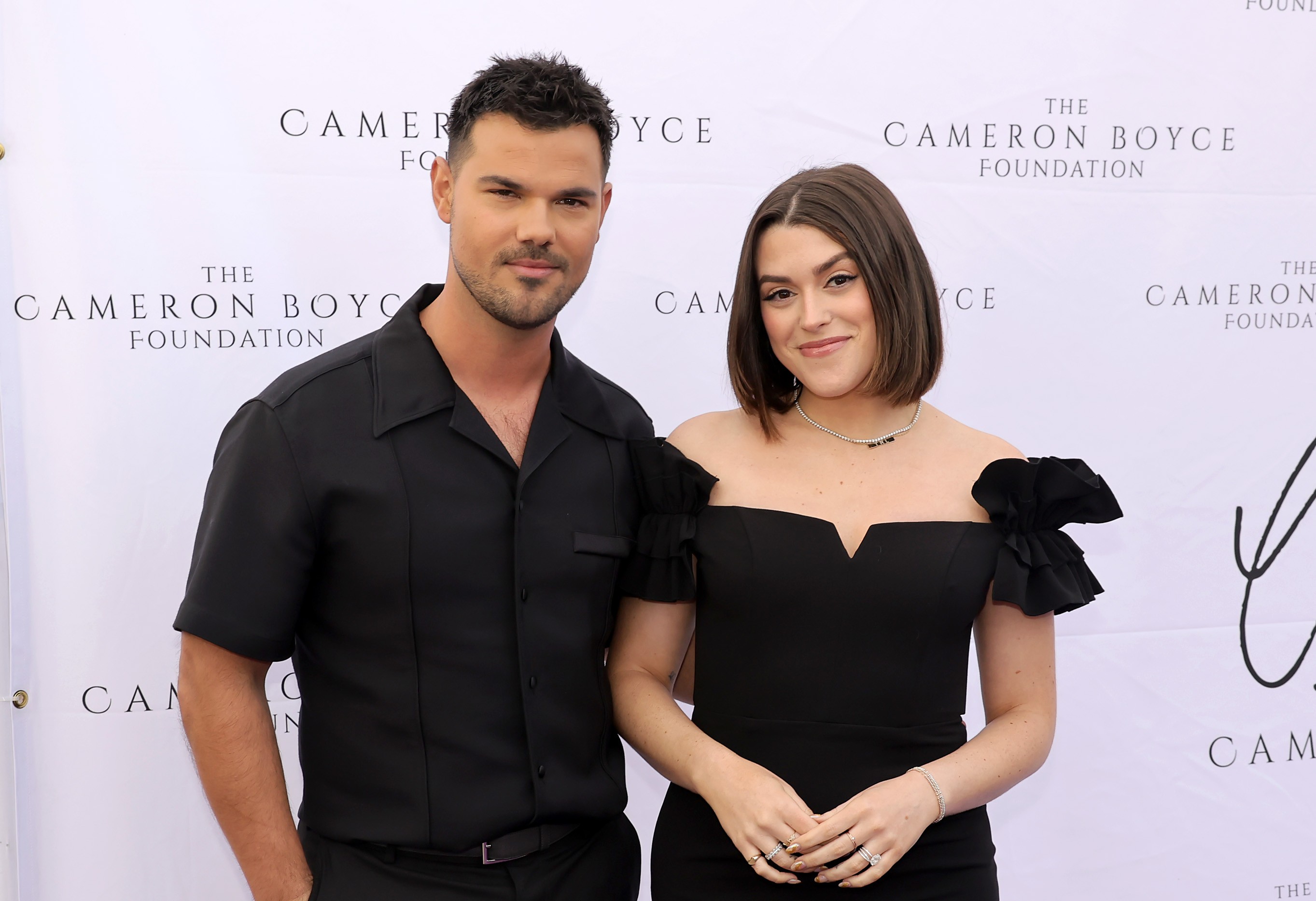 Taylor Lautner e Taylor Dome posam sorridentes, ele de camisa preta de manga curta e ela de vestido preto tomara que caia com detalhes nos ombros, em frente a um banner branco com o logo The Cameron Boyce Foundation