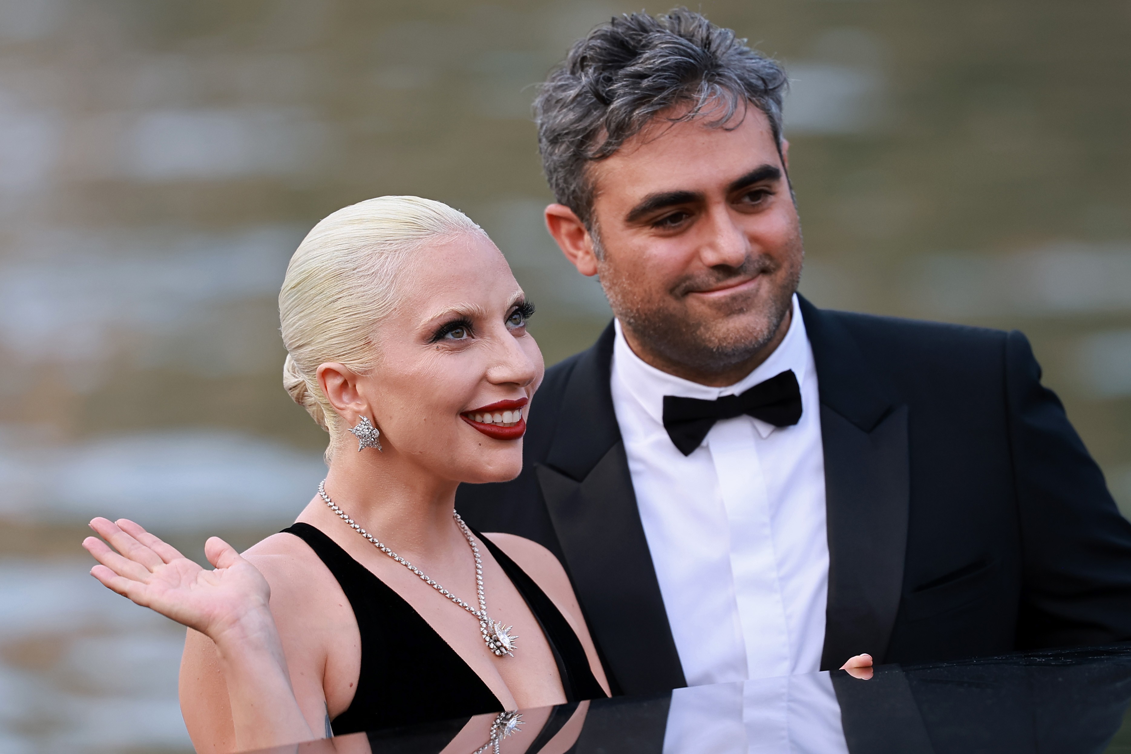 Lady Gaga, loira platinada com batom vermelho e colar brilhante, acena sorrindo ao lado de um homem de terno e gravata borboleta preta, ambos em um evento formal