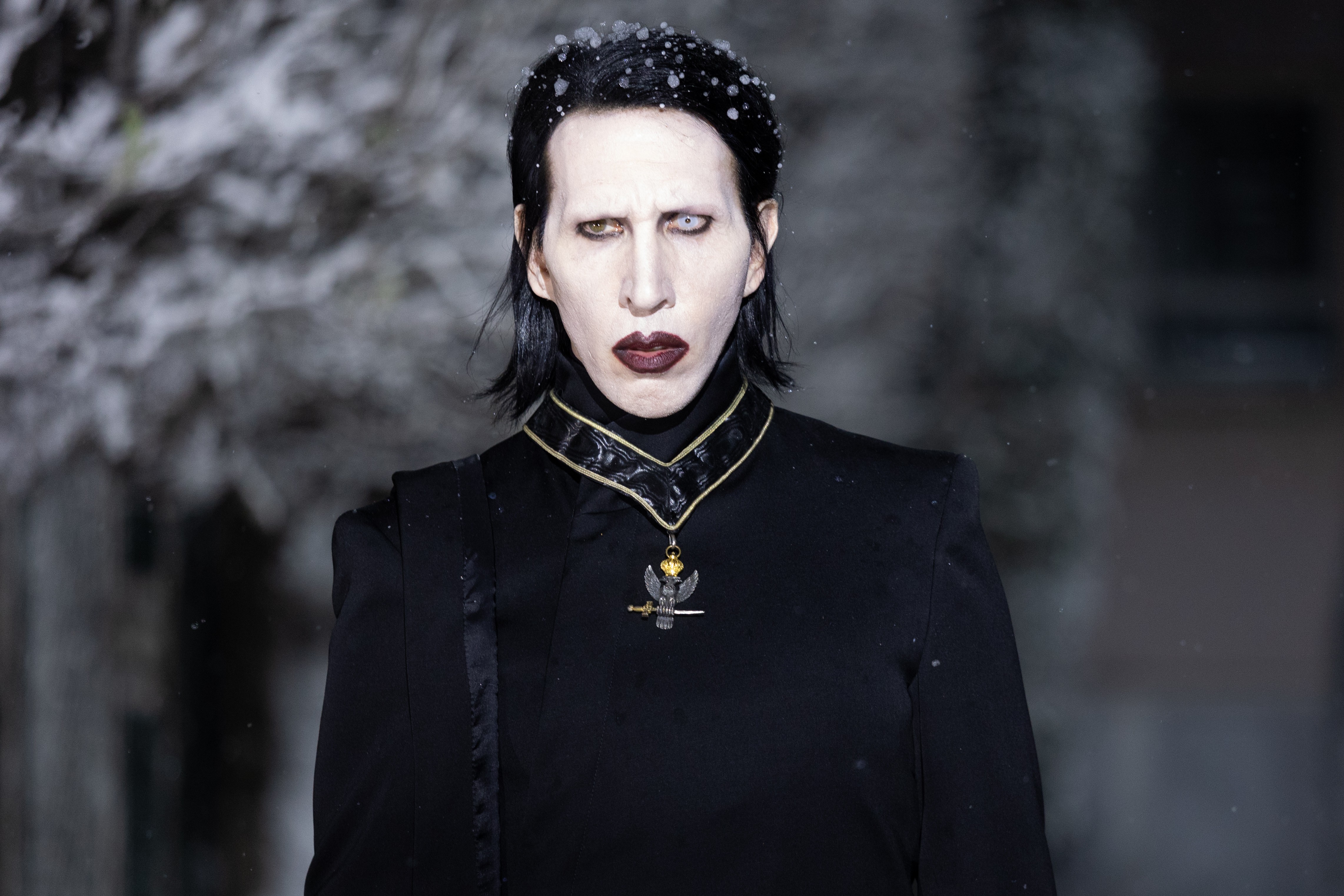 Marilyn Manson com maquiagem pálida, lábios escuros e olhos heterocromáticos, usando um casaco preto com gola alta e um medalhão de águia. Neve cai sobre seus cabelos escuros, em um cenário invernal