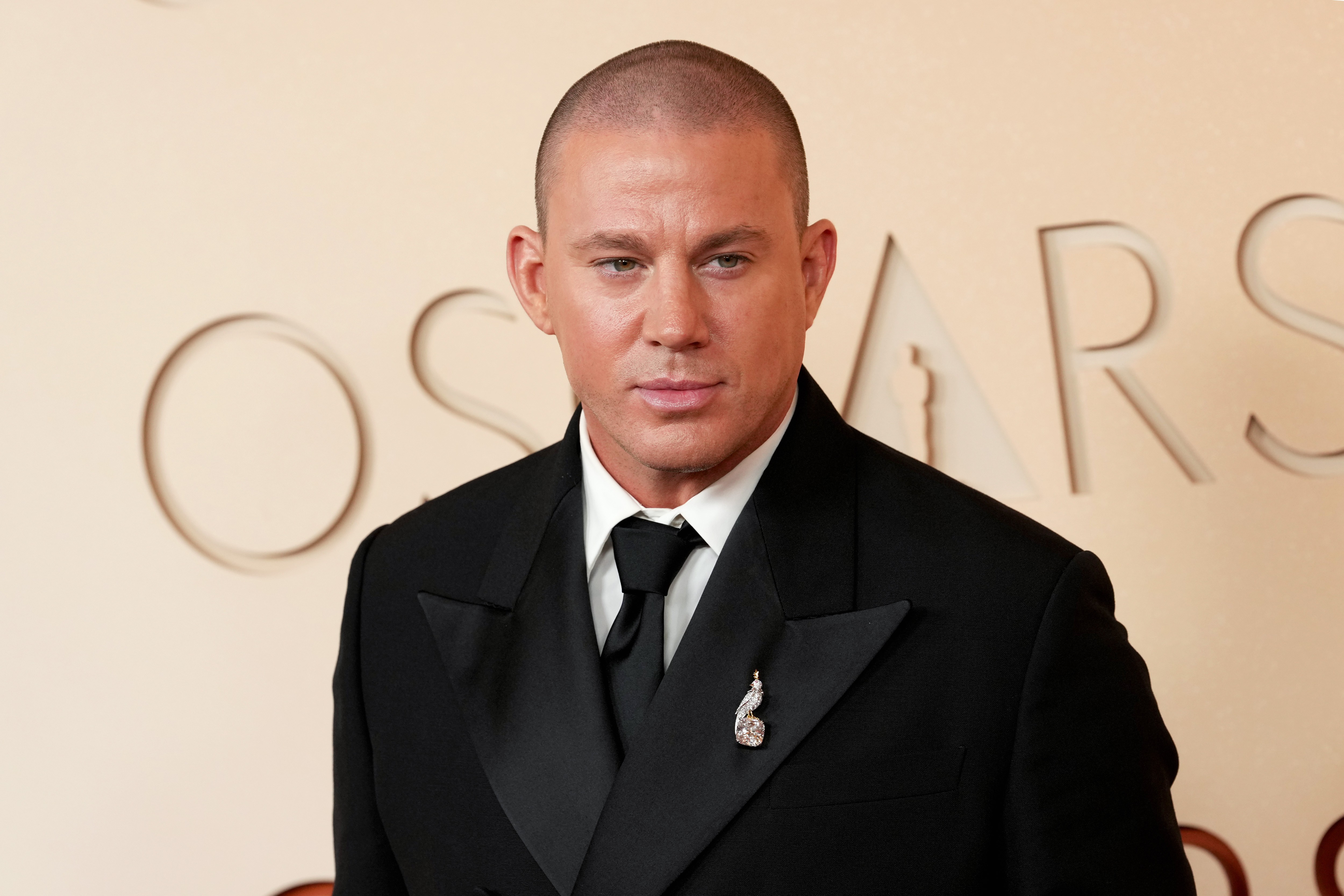 Channing Tatum, com cabelo raspado e expressão séria, veste um smoking preto com lapela de cetim, camisa branca e gravata preta. Um broche prateado adorna a lapela. Ao fundo, letras douradas OSCARS em relevo sobre uma parede bege