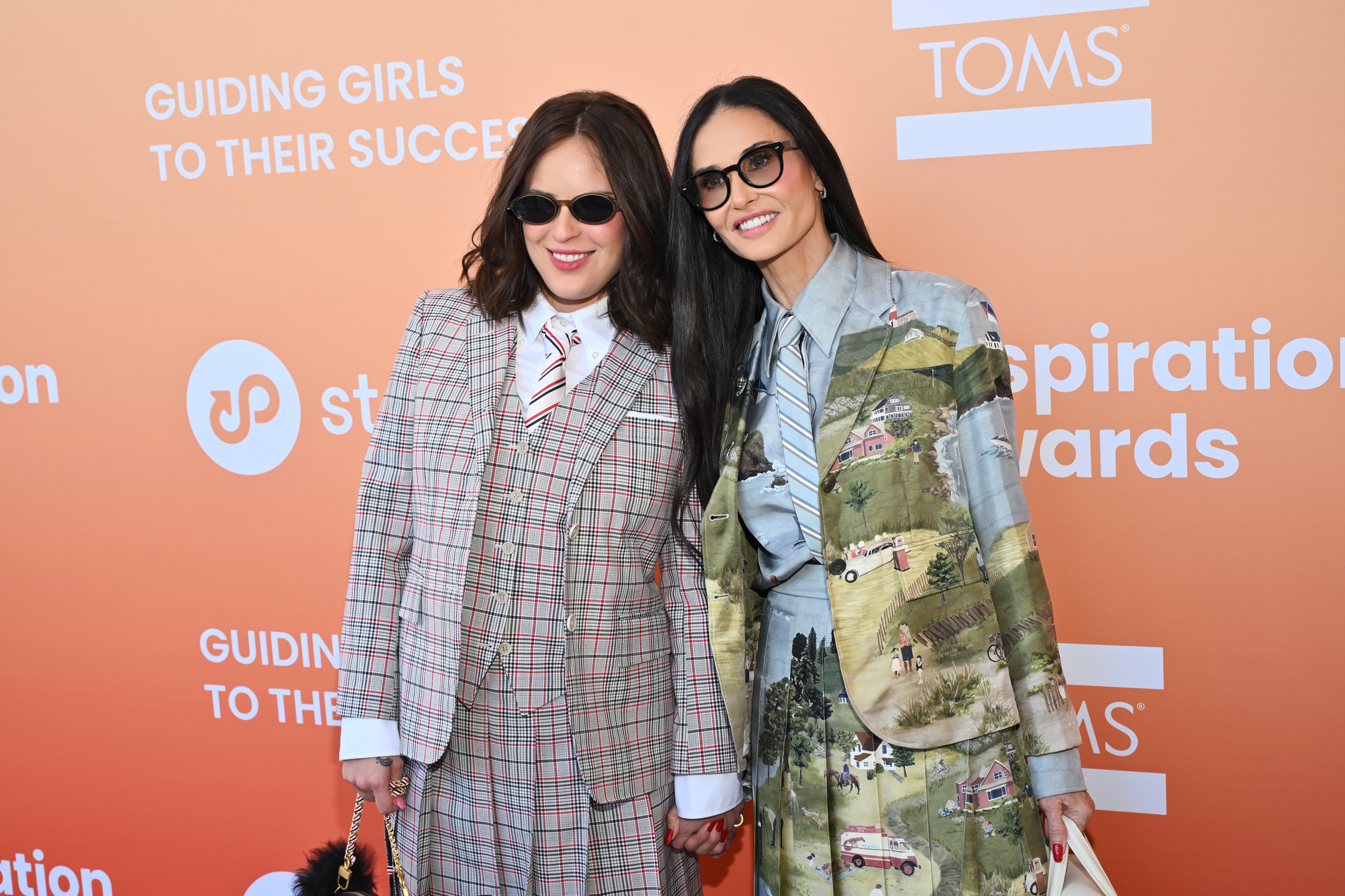 Duas mulheres sorridentes, Rumer Willis e Demi Moore, posam de mãos dadas em um evento. Rumer usa um terno xadrez com gravata listrada e óculos escuros ovais. Demi veste um terno estampado com paisagens rurais, gravata azul listrada e óculos escuros retangulares. O fundo laranja exibe logotipos e texto GUIDING GIRLS TO THEIR SUCCESS