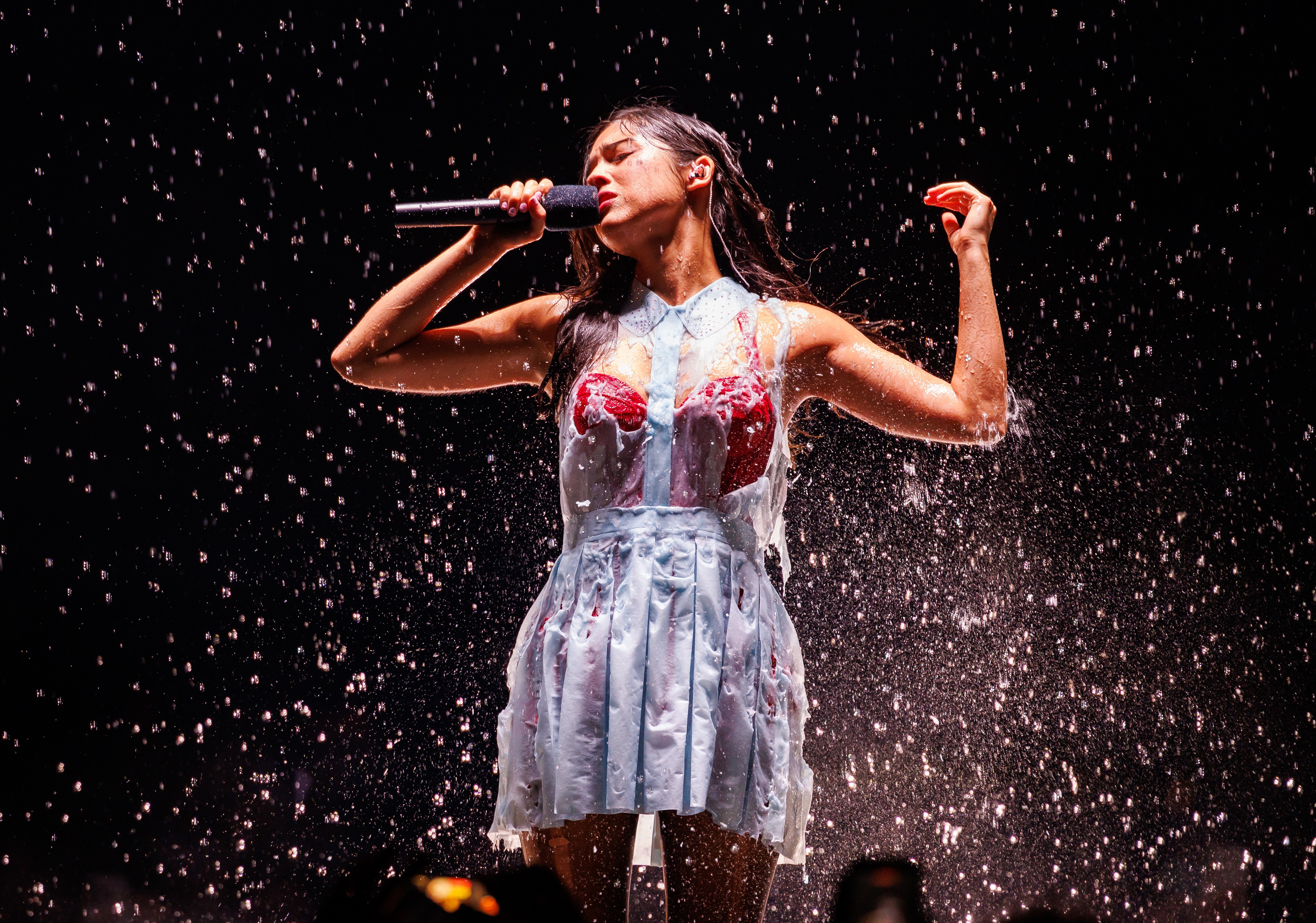 Olivia Rodrigo cantando em um palco escuro, encharcada de água e com um líquido vermelho escorrendo pelo vestido azul claro e sutiã vermelho, com os braços levantados e segurando um microfone