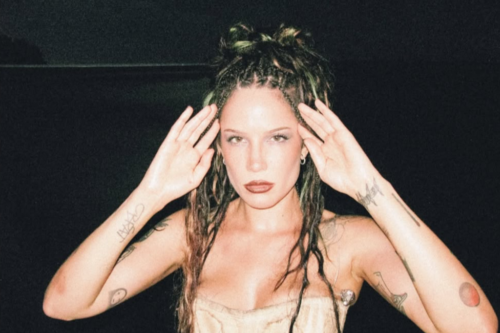 Cantora Halsey com cabelo trançado preso no alto e solto, maquiagem escura nos olhos e lábios, usando um top claro, com as mãos levantadas ao lado da cabeça, em fundo escuro