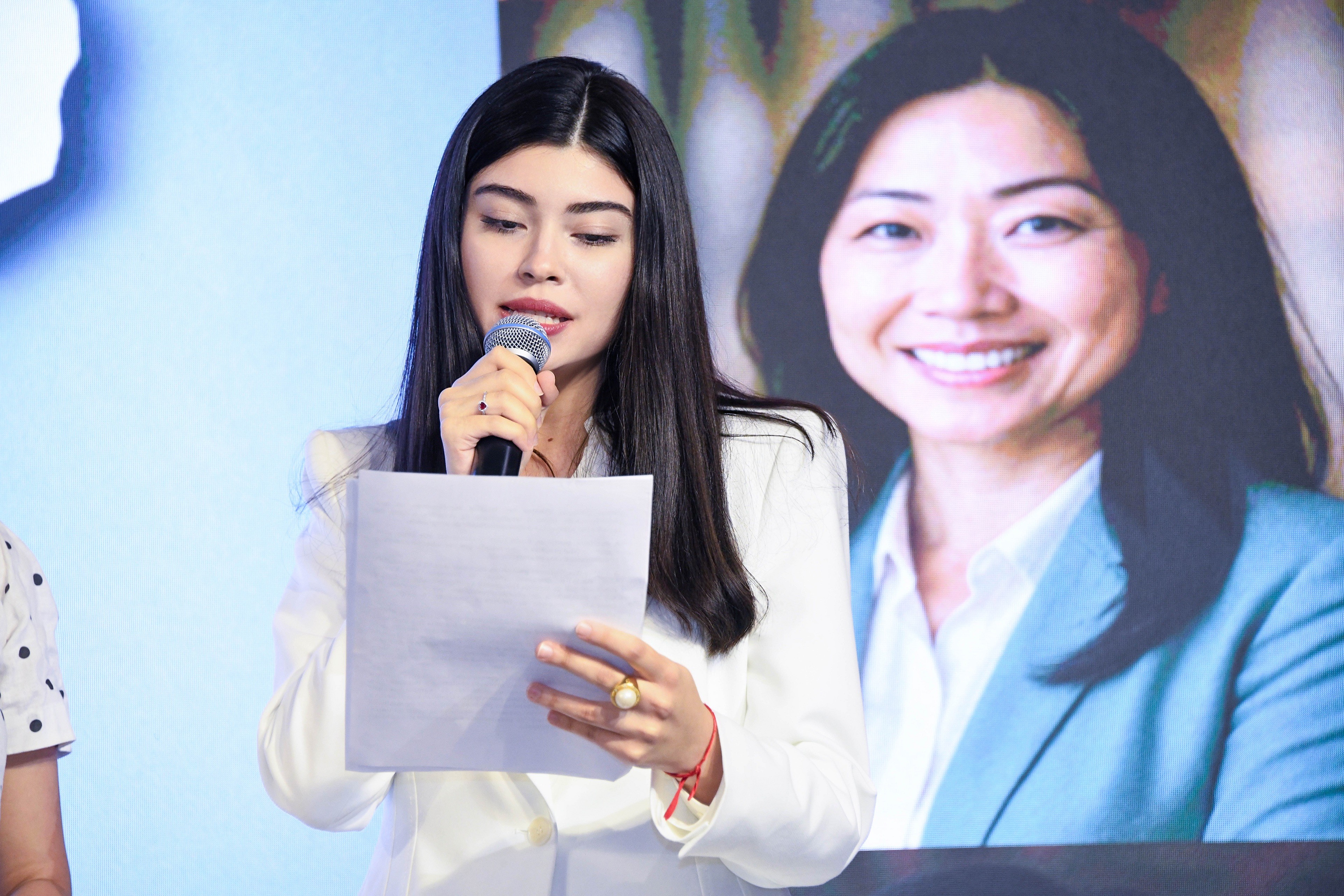 Mulher de cabelos longos e escuros, vestindo blazer branco, fala ao microfone segurando papéis. Ao fundo, uma projeção mostra o rosto sorridente de outra mulher asiática