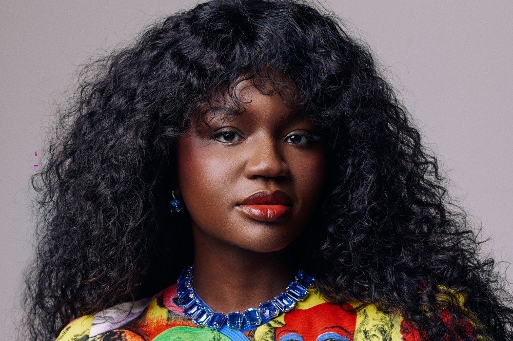 Mulher negra com cabelo crespo volumoso, batom laranja e colar azul vibrante, vestindo blusa colorida, olhando para frente com expressão séria