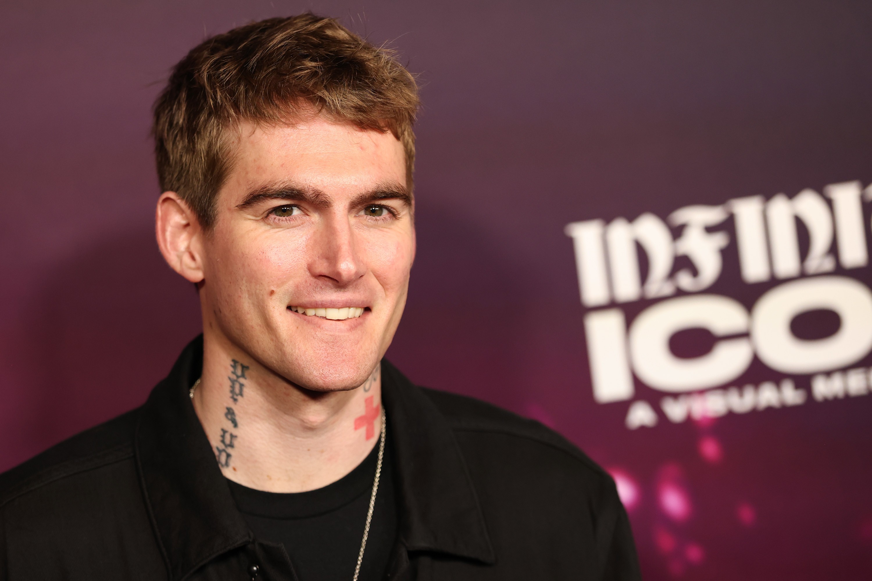 Homem branco de cabelo castanho claro, sorrindo, com tatuagens no pescoço e uma cruz vermelha. Veste camisa preta, em frente a um fundo roxo com o texto INFINITE ICON