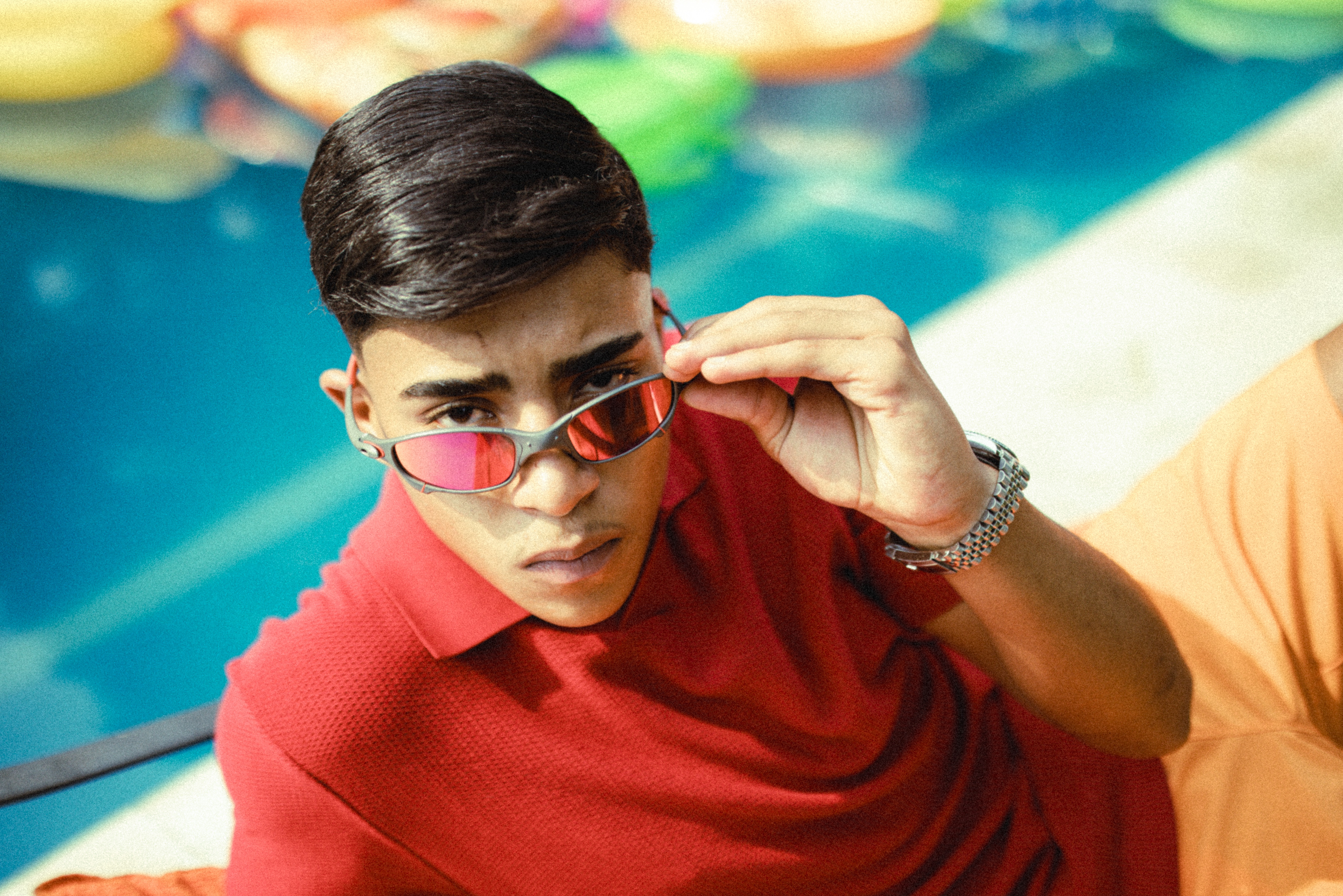Jovem de pele morena e cabelo escuro, vestindo camisa polo vermelha, abaixa os óculos de sol com lentes espelhadas vermelhas, olhando fixamente para frente. Ele está à beira de uma piscina azul, com boias coloridas ao fundo