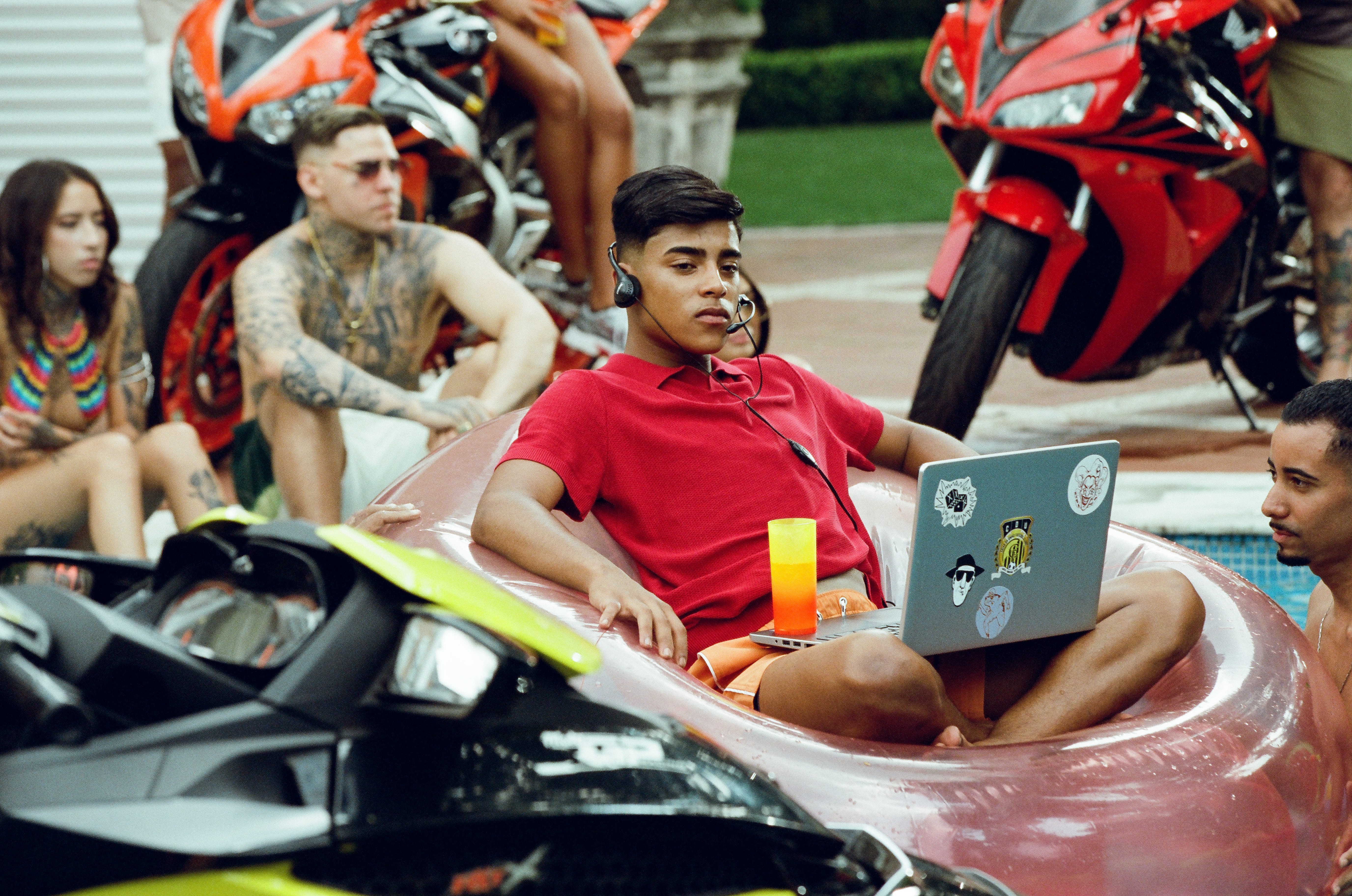 Jovem de camisa polo vermelha e shorts laranja sentado em boia inflável na piscina, usando headset e notebook com adesivos. Ao redor, pessoas tatuadas, motos esportivas e um jet ski, sugerindo um ambiente de lazer e ostentação