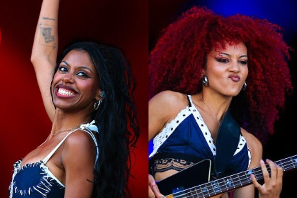 Duas mulheres negras em close-up. À esquerda, uma mulher de cabelos longos e pretos sorri, braço erguido. À direita, uma mulher de cabelo afro vermelho toca baixo, fazendo bico