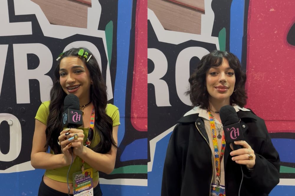 Duas jovens sorrindo para a câmera, segurando microfones. A da esquerda tem cabelos longos, usa blusas verde-limão e presilhas verdes. A da direita tem cabelos curtos, veste jaqueta preta e blusa bege