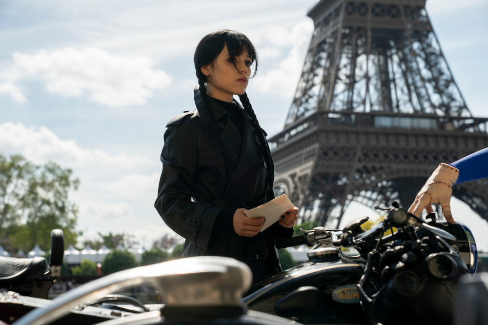 Mulher com tranças e casaco preto segura um envelope, ao lado de uma moto, com a Torre Eiffel ao fundo sob céu azul
