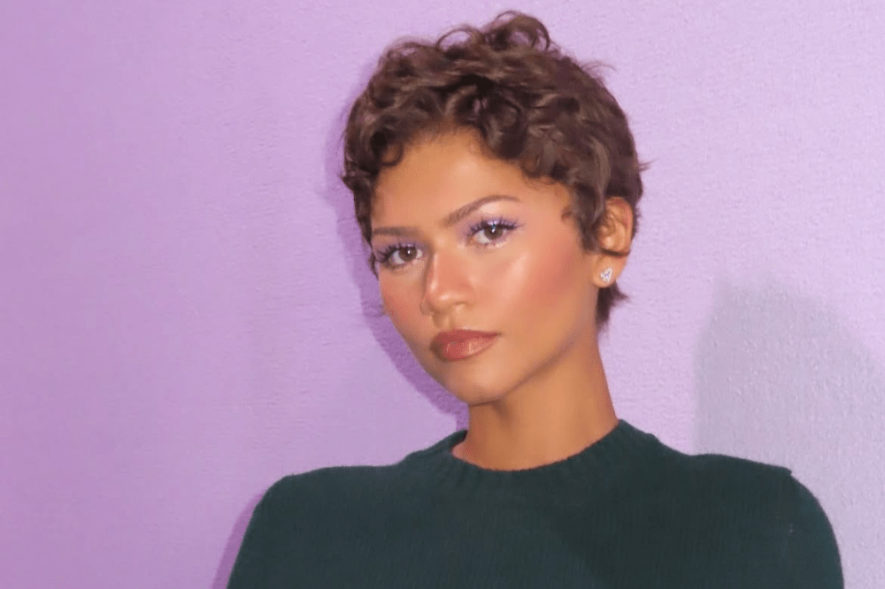 Mulher jovem de pele morena, cabelo castanho curto e encaracolado, maquiagem roxa nos olhos e batom nude, usando blusa verde escura e brinco de borboleta, posa com expressão séria contra uma parede lilás