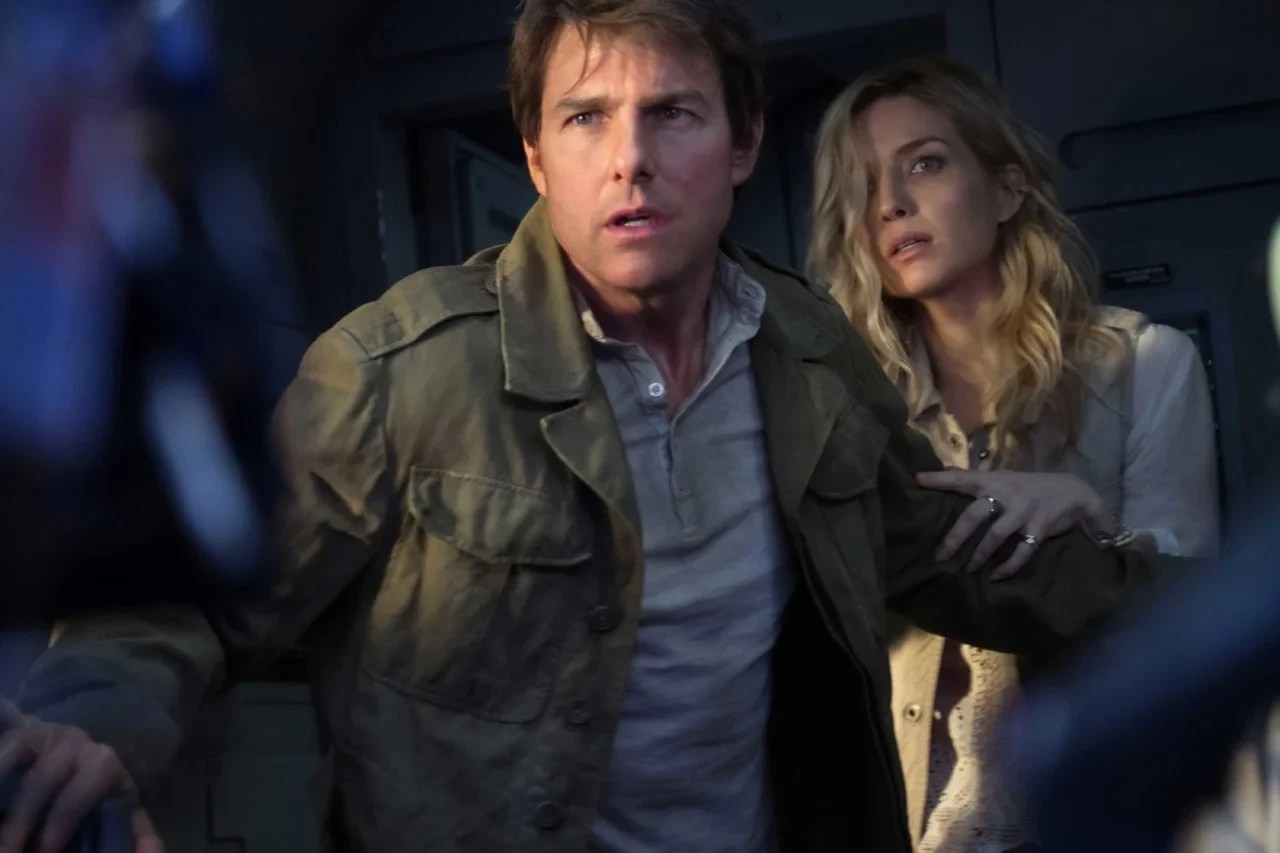 Tom Cruise, com jaqueta verde-oliva e camisa azul-clara, olha assustado para cima, boca entreaberta. Ao seu lado, uma mulher loira com blusa clara e casaco bege, também com expressão de pavor, segura o braço dele. Ambos estão em um ambiente escuro, possivelmente dentro de um veículo ou estrutura, com luzes azuis e brancas ao fundo
