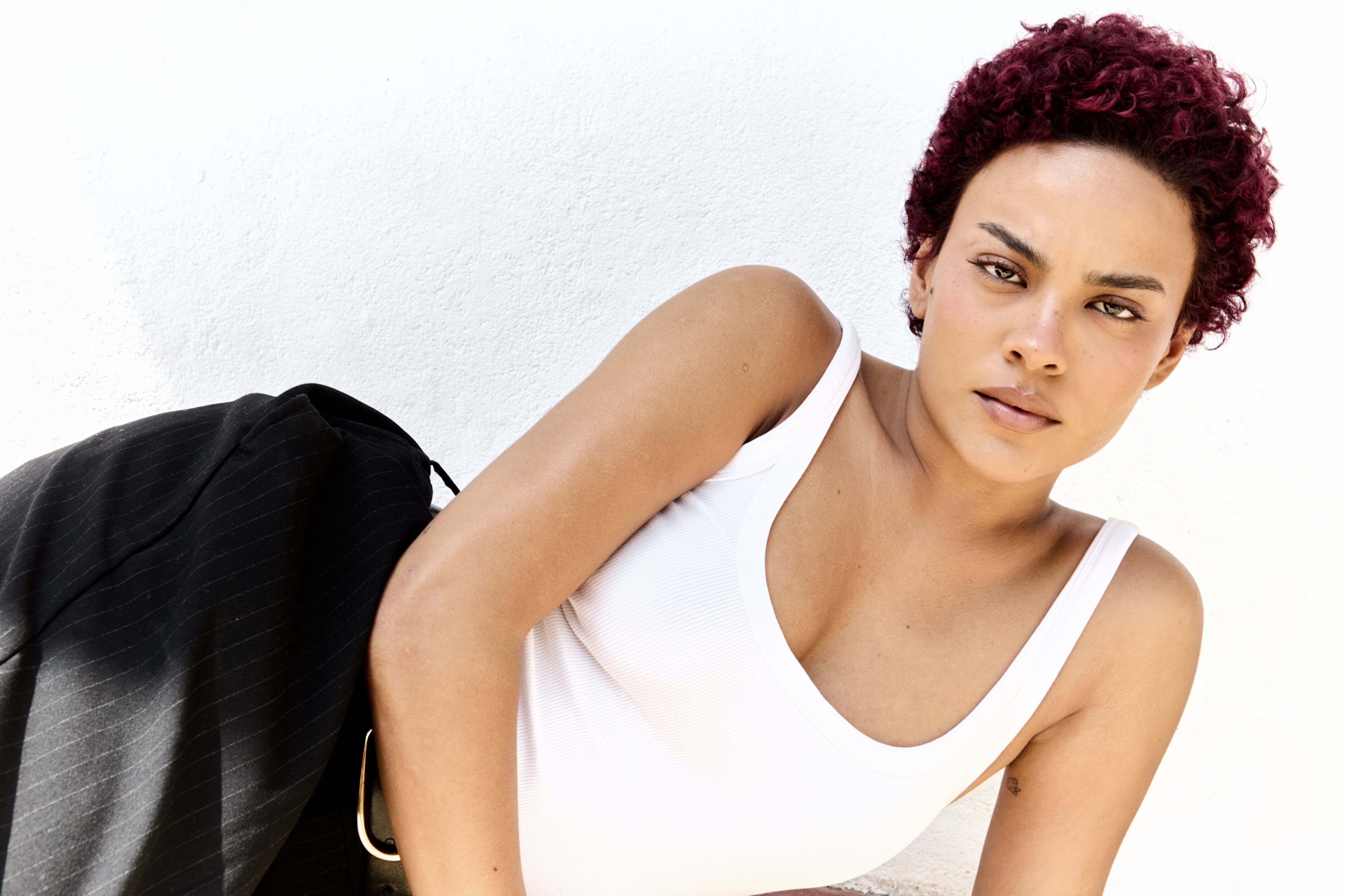 Mulher de pele morena clara, cabelo crespo curto e ruivo, vestindo regata branca e calça preta risca de giz, olhando diretamente para frente com expressão séria, encostada em uma parede branca