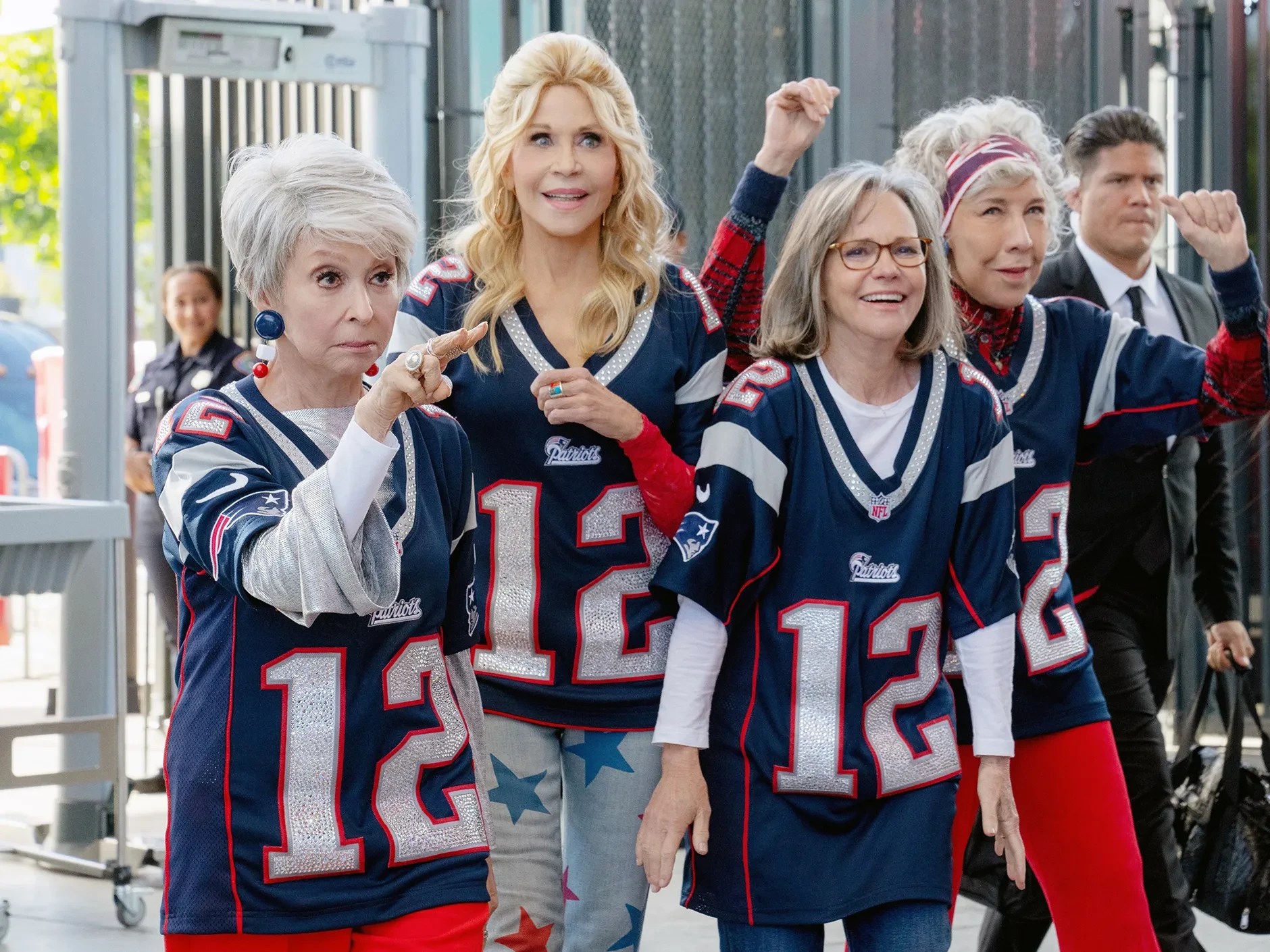 Quatro mulheres idosas, sorrindo e animadas, vestem camisetas azuis do time Patriots com o número 12 em glitter. Uma delas aponta para frente, enquanto outra levanta o braço em celebração. Ao fundo, um homem de terno e uma mulher de uniforme de segurança observam.