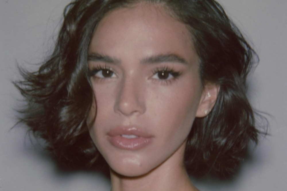 Rosto de uma mulher jovem com cabelo curto e ondulado, castanho escuro, olhando diretamente para frente. Ela tem olhos grandes, maquiagem leve nos olhos e lábios rosados e cheios. A pele é clara e o fundo é um cinza claro