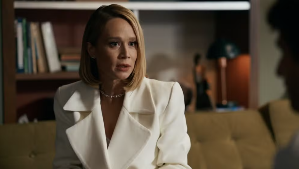 Mulher loira de cabelo curto e liso, vestindo blazer branco e colar prateado, sentada em um sofá mostarda, conversando com alguém à direita. Ao fundo, uma estante com livros e uma luminária
