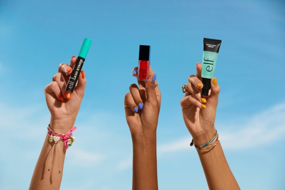 Três mãos erguidas contra um céu azul claro, cada uma segurando um produto de maquiagem da marca e.l.f. Cosmetics. Da esquerda para a direita: uma mão com unhas laranja e pulseiras coloridas segurando um rímel preto e verde-água; uma mão com unhas lilás segurando um batom líquido vermelho; e uma mão com unhas amarelas e pulseiras finas segurando um primer facial em tubo preto e verde-água