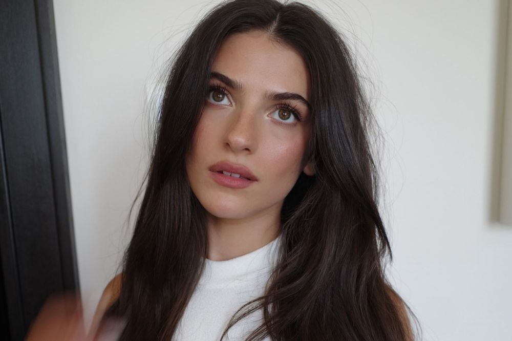Jovem mulher de cabelos castanhos escuros e ondulados, olhando para cima com expressão pensativa, usando blusa branca, em fundo claro