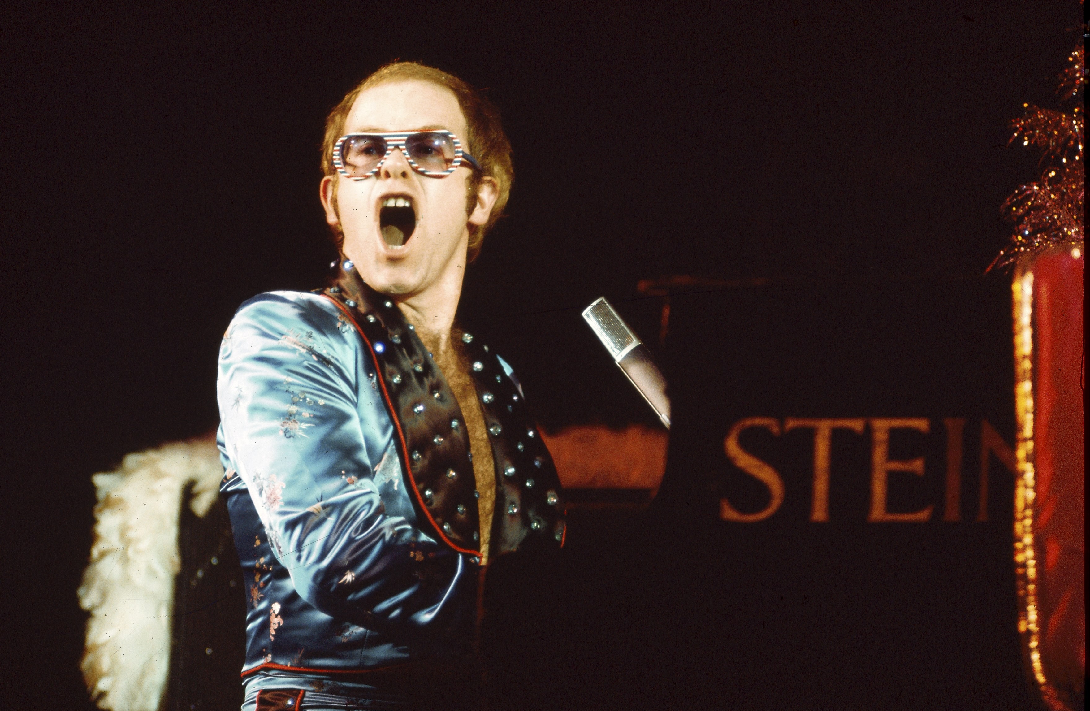 Elton John, com óculos de sol listrados e jaqueta azul brilhante, canta com a boca aberta e expressiva, em frente a um piano escuro com a palavra STEIN visível