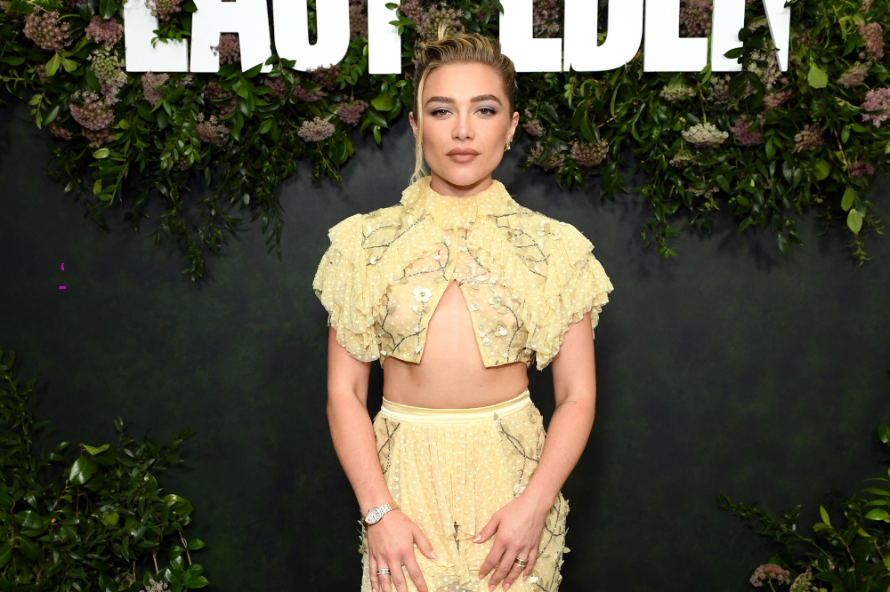 A atriz Florence Pugh posa sorrindo, com cabelo loiro preso, maquiagem verde nos olhos e batom nude. Ela veste um conjunto amarelo claro bordado, com top cropped de gola alta e saia longa, exibindo a barriga. Ao fundo, uma parede escura decorada com folhagens verdes e flores secas, e letras brancas de um letreiro