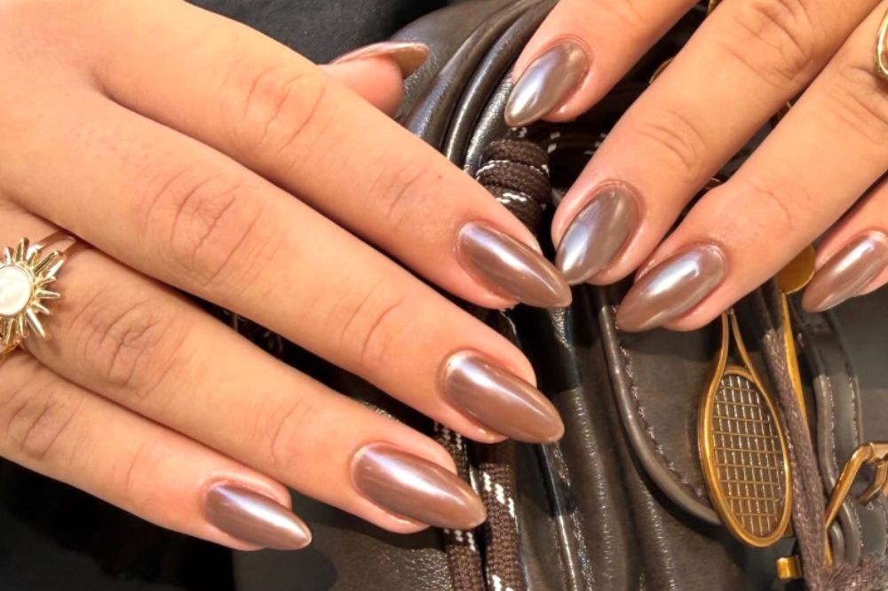 Mãos de uma pessoa com pele morena e unhas amendoadas esmaltadas em marrom perolado, segurando uma bolsa escura com um pingente de raquete de tênis dourada. Um anel dourado com um sol e centro branco está no dedo anelar esquerdo