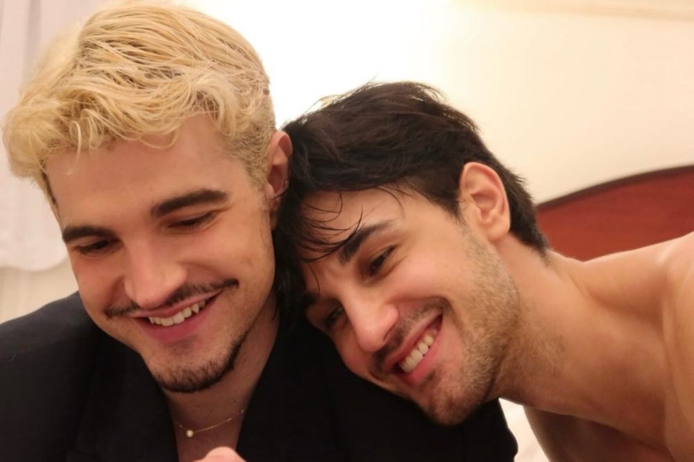 Dois homens jovens sorrindo, um com cabelo loiro e bigode, o outro com cabelo escuro e barba rala, encostando a cabeça no ombro do primeiro