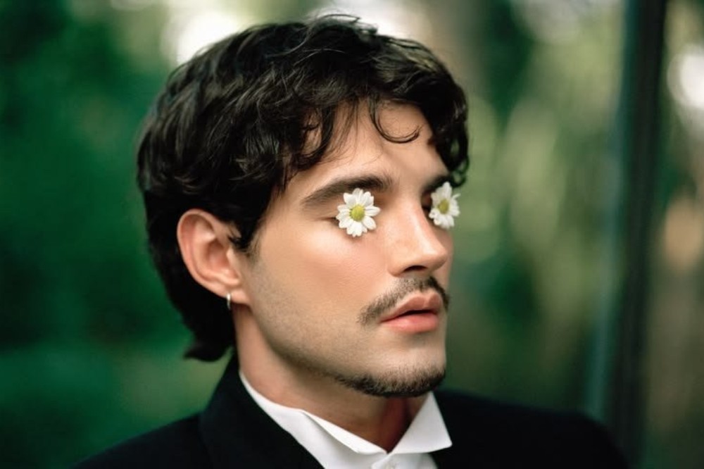 Homem jovem, de pele clara, cabelo escuro e ondulado, com bigode e barba rala, usando terno preto e camisa branca. Duas margaridas brancas cobrem seus olhos, e ele usa um brinco de argola prateado na orelha esquerda. O fundo é desfocado em tons de verde