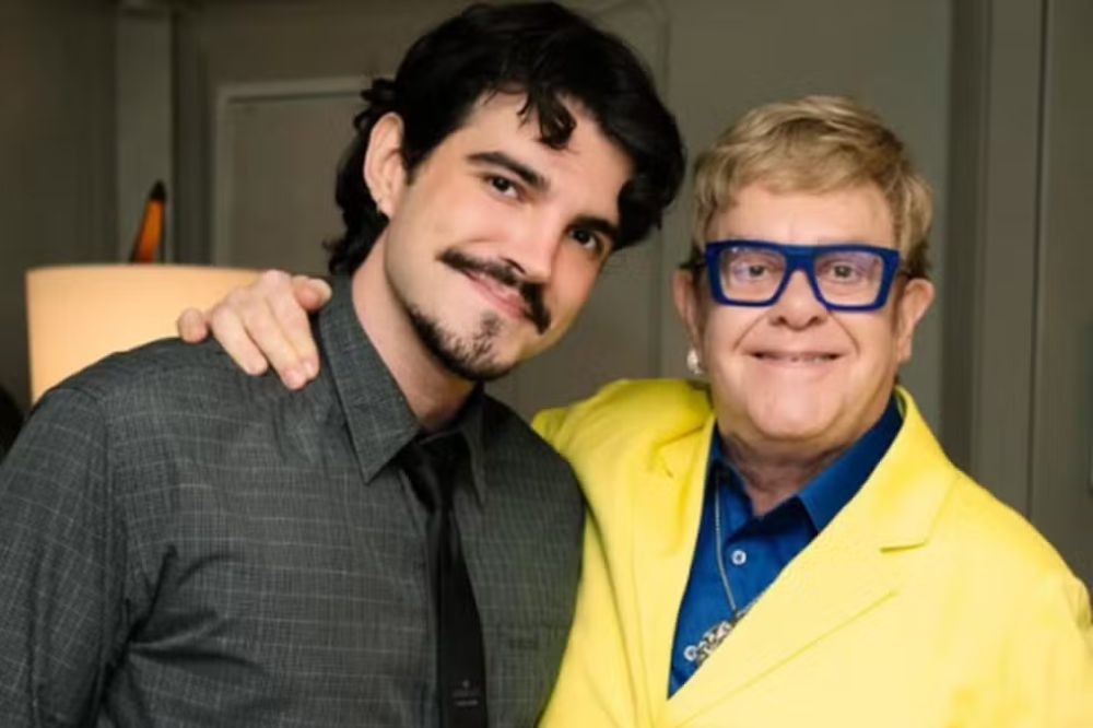 Um homem jovem de bigode e barba rala, com camisa xadrez escura e gravata preta, abraça Elton John, que veste blazer amarelo, camisa azul e óculos azuis, ambos sorrindo para a câmera
