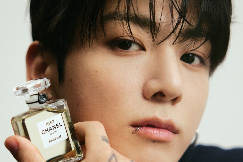 Close-up de um homem jovem com cabelo escuro e úmido, segurando um frasco de perfume Chanel 1957. Ele tem um piercing prateado no lábio inferior e tatuagens nos dedos, olhando diretamente para a câmera com expressão séria