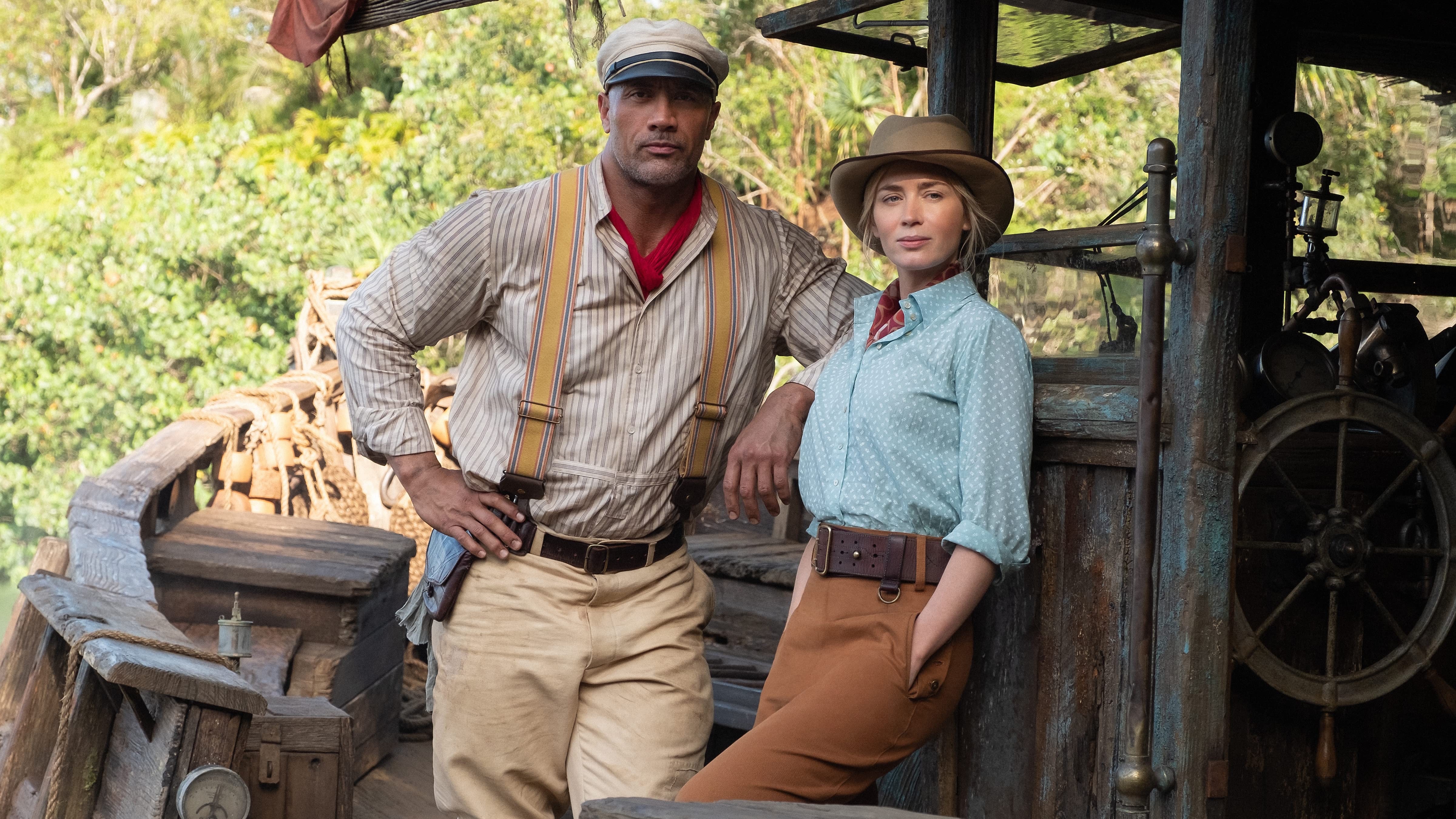 Dwayne The Rock Johnson e Emily Blunt posam em um barco rústico. Ele veste camisa listrada, suspensórios e boné, com um lenço vermelho. Ela usa camisa azul clara de bolinhas, calça marrom e chapéu. Ambos olham para frente com expressões sérias, em um cenário de selva