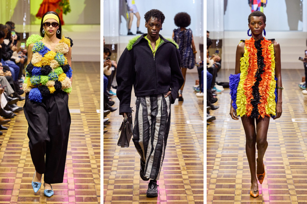 Três modelos desfilam em passarela de madeira. À esquerda, mulher com top de pompons coloridos e calça preta. Ao centro, homem com jaqueta preta, camisa verde-limão e calça listrada. À direita, mulher com vestido curto de babados coloridos e brincos azuis