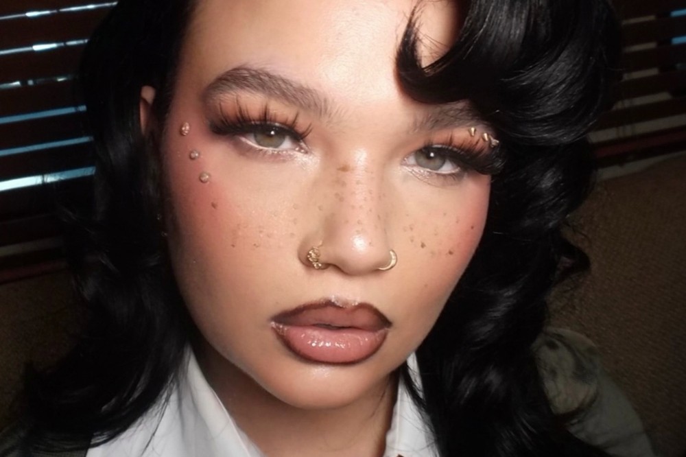 Mulher jovem com cabelo preto ondulado, maquiagem elaborada com delineador branco, cílios postiços volumosos, sardas e blush rosado. Ela tem piercings dourados no nariz e pedrinhas brilhantes no rosto, perto dos olhos. Seus lábios estão contornados em marrom e preenchidos com gloss nude.