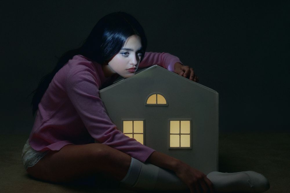 Jovem de cabelos escuros e lisos, vestindo blusa rosa e shorts brancos, sentada no chão escuro, abraça uma casa de brinquedo iluminada por dentro, com janelas brilhando em amarelo, e olha para a frente com expressão séria