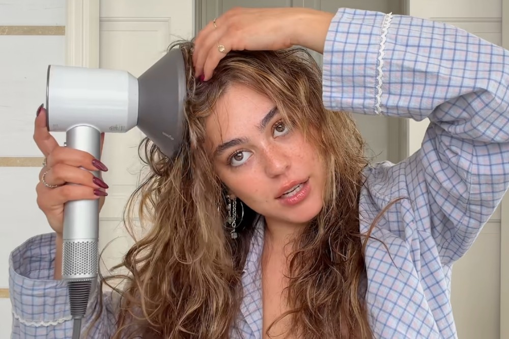 Mulher jovem com cabelos castanhos ondulados e molhados, vestindo pijama xadrez azul e branco, secando a raiz do cabelo com um secador branco e cinza com difusor, olhando para frente