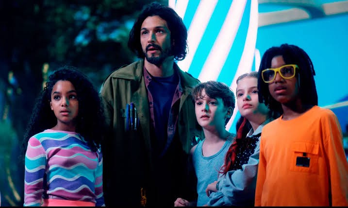 Um homem barbudo de cabelo comprido e quatro crianças, duas meninas e dois meninos, olham para cima com expressões de surpresa. O homem veste jaqueta verde-oliva, uma menina tem cabelo cacheado e blusa listrada colorida, outra menina tem cabelo ruivo e blusa escura, um menino veste regata azul e o outro, de óculos amarelos, veste blusa laranja. O fundo é escuro com elementos azuis e brancos
