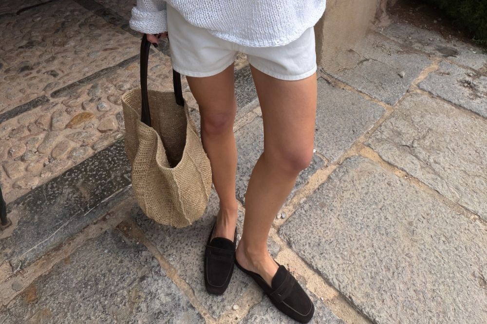 Pernas de uma pessoa em shorts brancos e suéter claro, segurando uma bolsa de palha com alças escuras, usando sapatos pretos tipo mule, caminhando sobre um piso de pedras irregulares