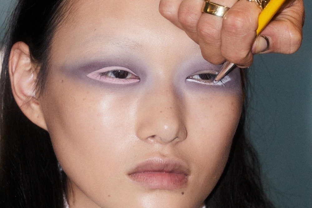 Close-up de uma pessoa com maquiagem artística. Um maquiador, com anéis dourados, aplica delineador branco na pálpebra inferior do olho direito. A modelo tem sombra roxa nas pálpebras e um delineado rosa na pálpebra inferior esquerda